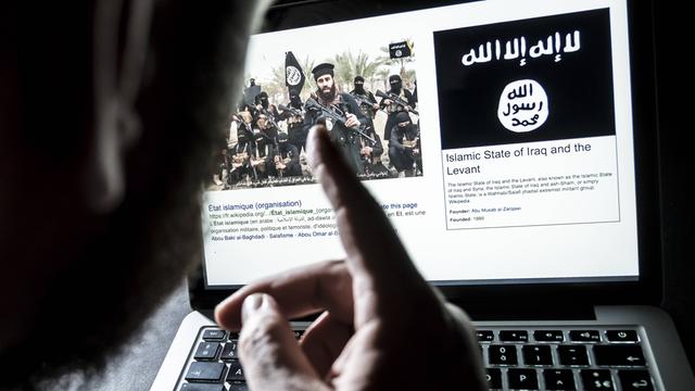 Ein Mann sitzt vor einem Computerbildschirm, auf dem eine offensichtlich islamistische Gruppe bärtiger Männer zu sehen ist, die Maschinengewehre in der Hand halten. Daneben sieht man die Flagge vom "Islamistischen Staat".