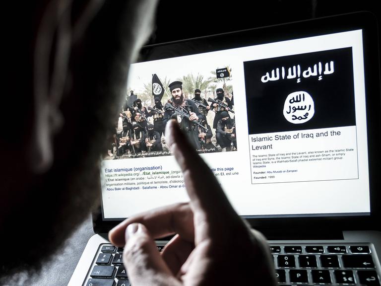 Ein Mann sitzt vor einem Computerbildschirm, auf dem eine offensichtlich islamistische Gruppe bärtiger Männer zu sehen ist, die Maschinengewehre in der Hand halten. Daneben sieht man die Flagge vom "Islamistischen Staat". Ein Mann sitzt vor einem Computerbildschirm, auf dem eine offensichtlich islamistische Gruppe bärtiger Männer zu sehen ist, die Maschinengewehre in der Hand halten. Daneben sieht man die Flagge vom "Islamistischen Staat".
