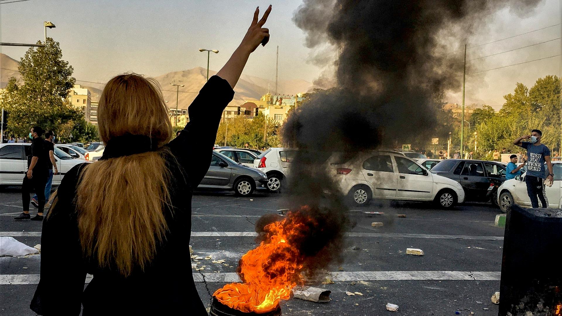 Proteste im Iran: Von hinten ist eine Frau zu sehen, sie trägt kein Kopftuch und reckt ihren Arm in die Höhe: Neben ihr brennt ein Feuer. 