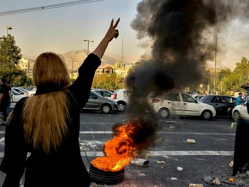 Proteste im Iran: Von hinten ist eine Frau zu sehen, sie trägt kein Kopftuch und reckt ihren Arm in die Höhe: Neben ihr brennt ein Feuer. 