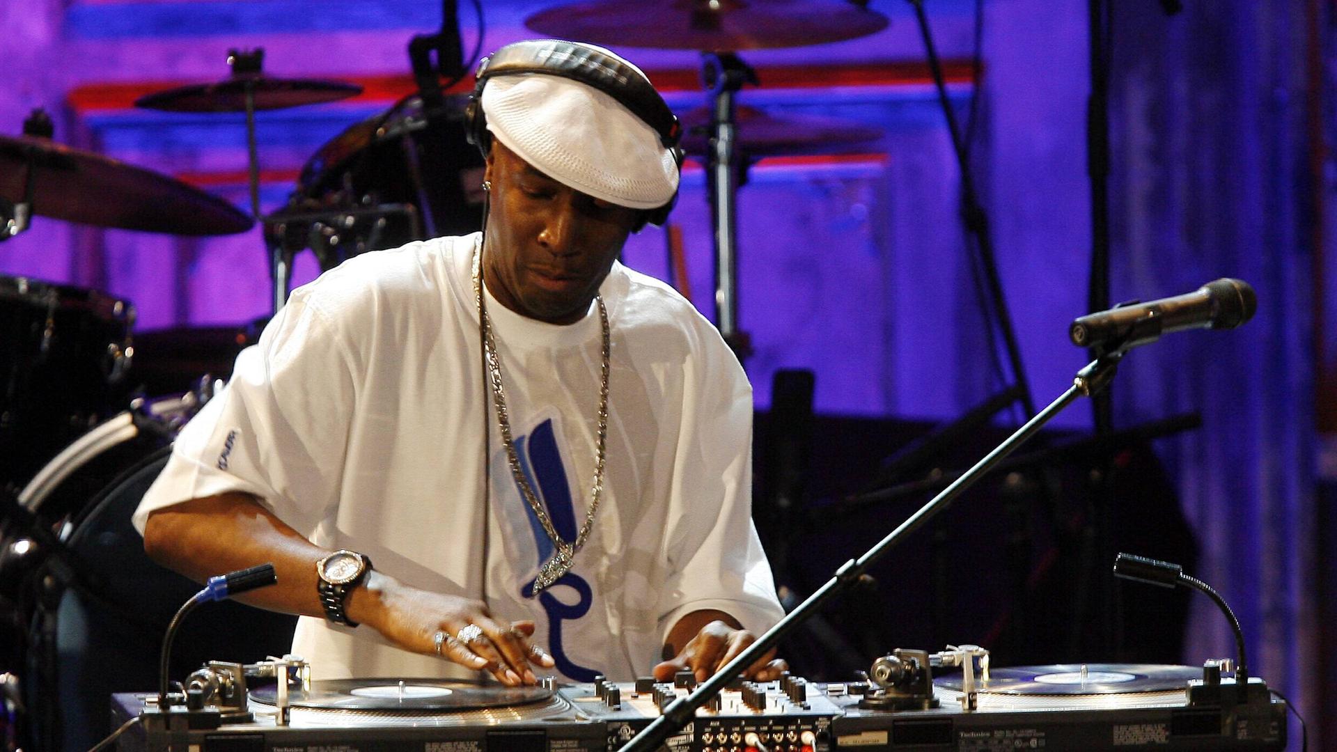 Ein Mann steht vor einem DJ Mischpult, die rechte Hand liegt auf der Platte. Er trägt Kopfhörer, ein weißes T-Shirt und eine weiße Schiebermütze.