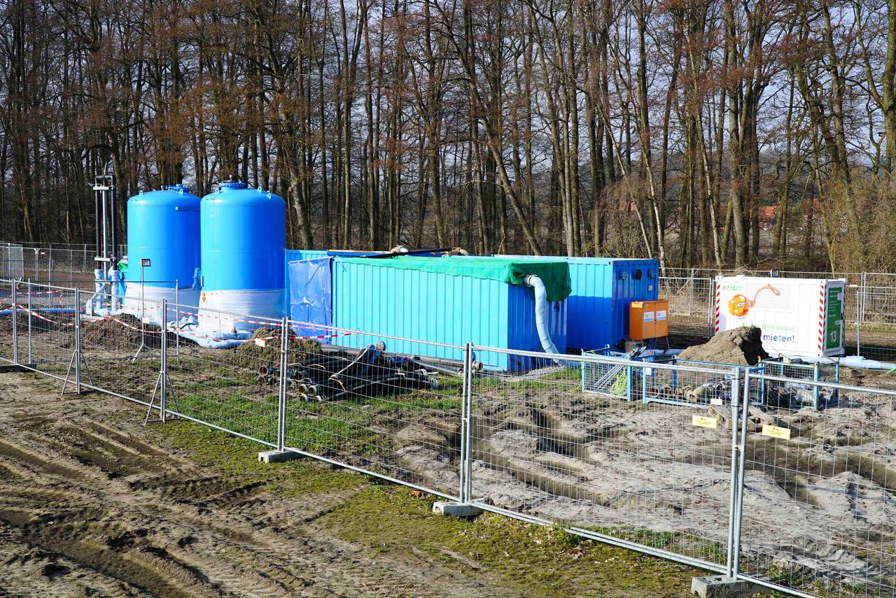 Auf einem eingezäunten Gelände in der Nähe einer Baumgruppe stehen Baumaschinen und runde blaue Tanks.