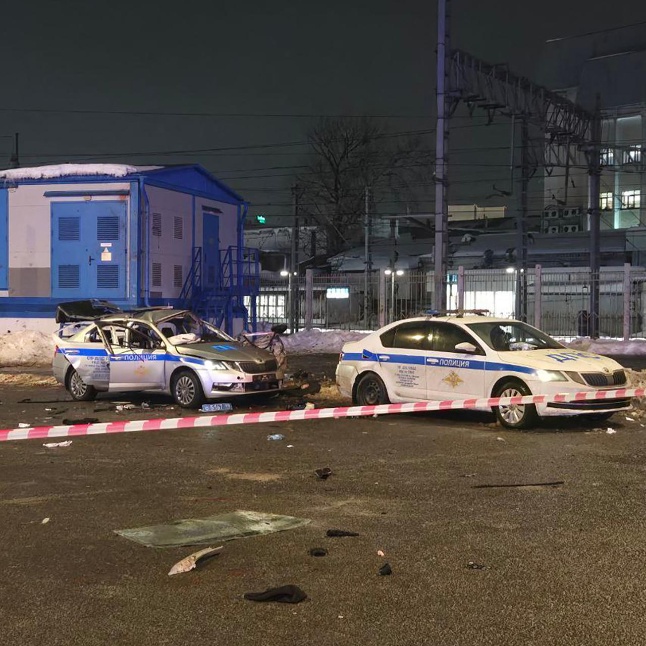 Das Bild zeigt ein zerstörtes Polizeiauto, davor ein intaktes Einsatzfahrzeug. Der Tatort ist mit einem rot-weißen Polizeiband abgeriegelt.