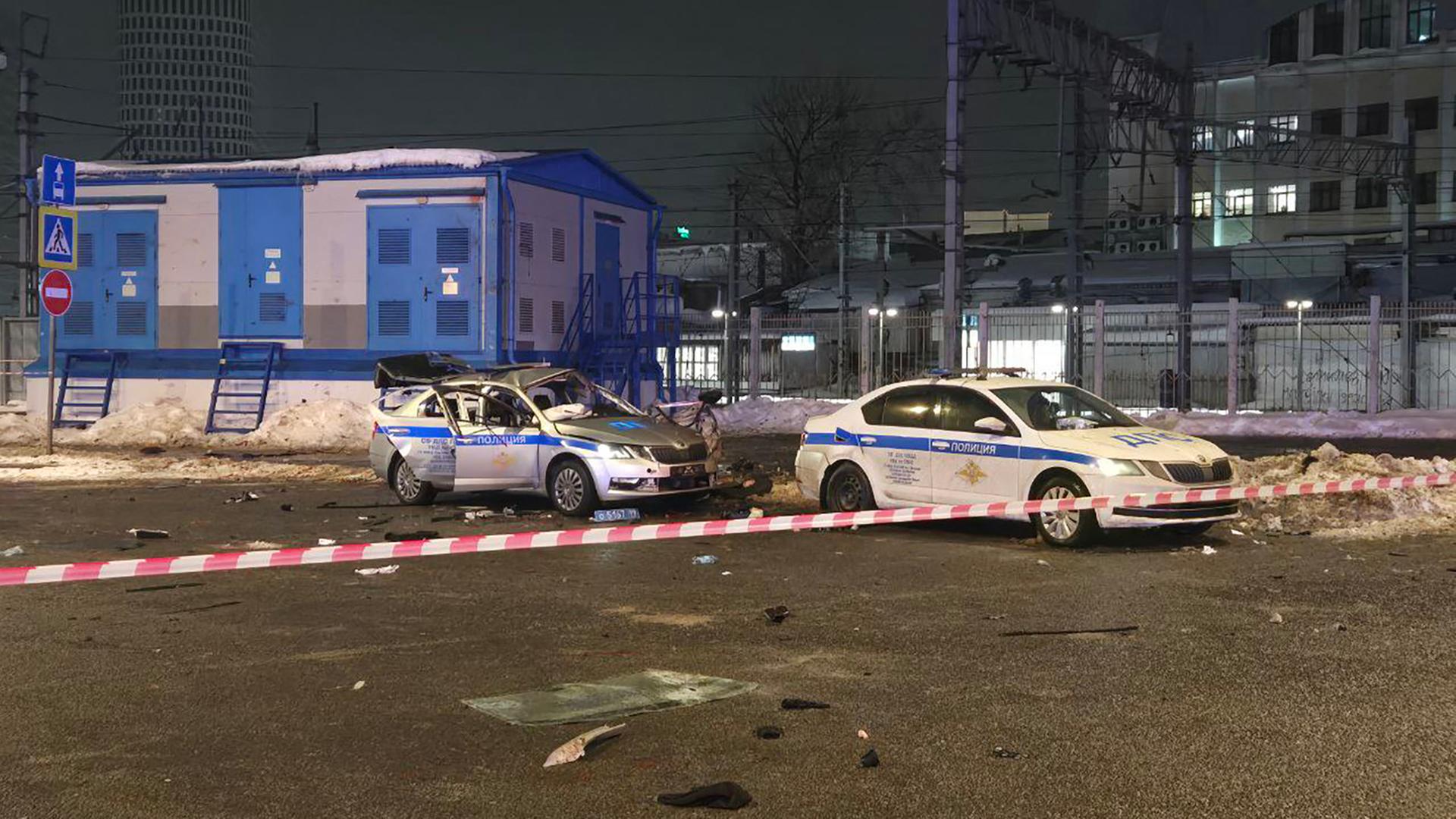 Das Bild zeigt ein zerstörtes Polizeiauto, davor ein intaktes Einsatzfahrzeug. Der Tatort ist mit einem rot-weißen Polizeiband abgeriegelt.
