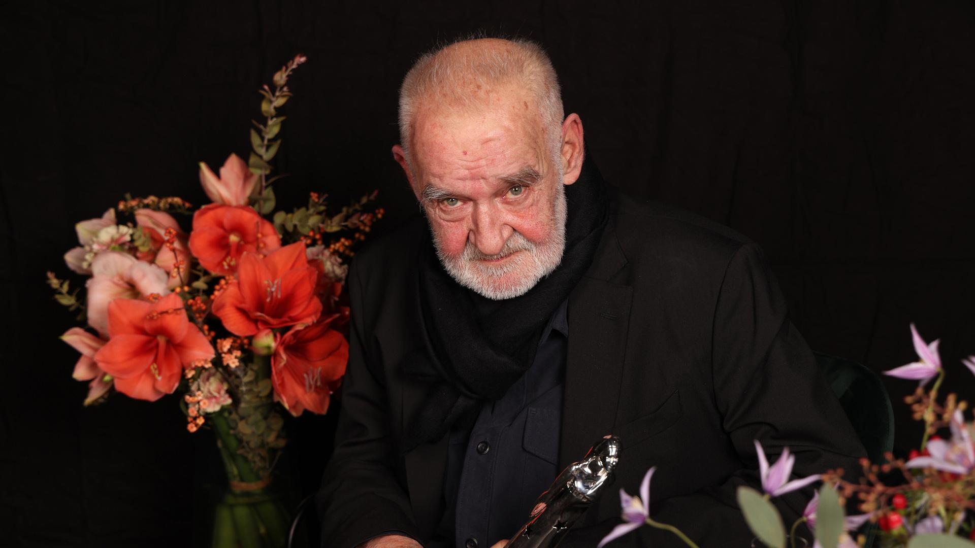 Béla Tarr sitzt neben Blumen vor dunklem Hintergrund.