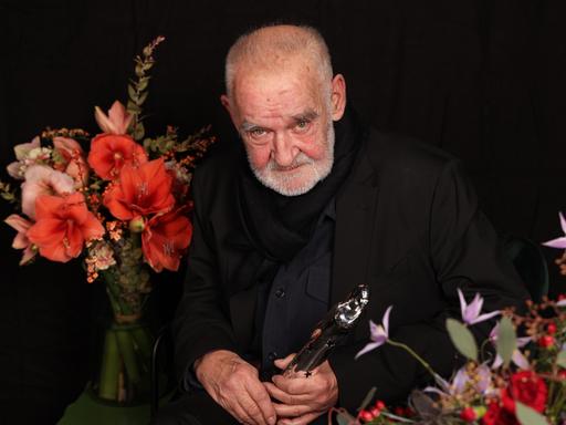 Béla Tarr sitzt neben Blumen vor dunklem Hintergrund.