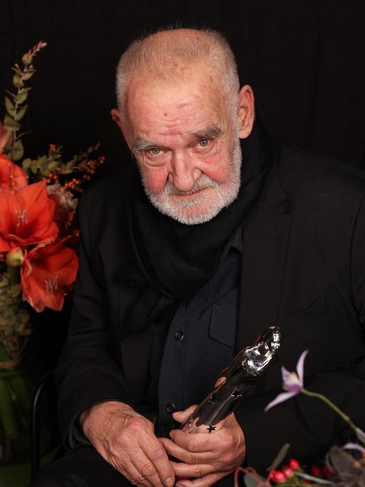 Bela Tarr sitzt neben Blumen vor dunklem Hintergrund.