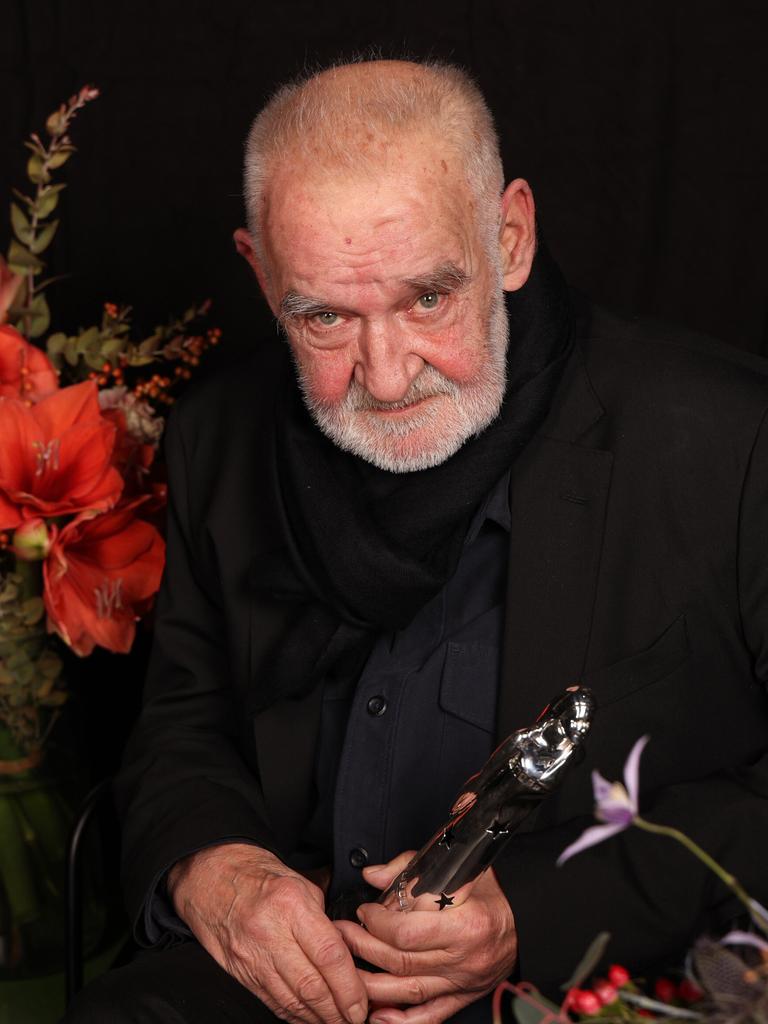 Béla Tarr sitzt neben Blumen vor dunklem Hintergrund.