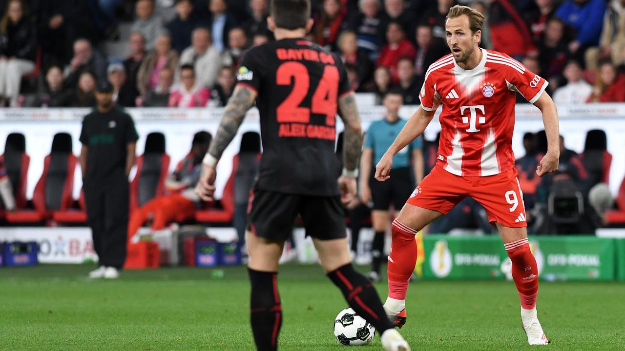Kane geht mit dem Ball am Fuß auf einen Gegenspieler zu. Kane trägt ein rotes Trikot, der Gegenspieler spielt in schwarz. Im Hintergrund steht Bayern-Trainer Vincent Kompany.