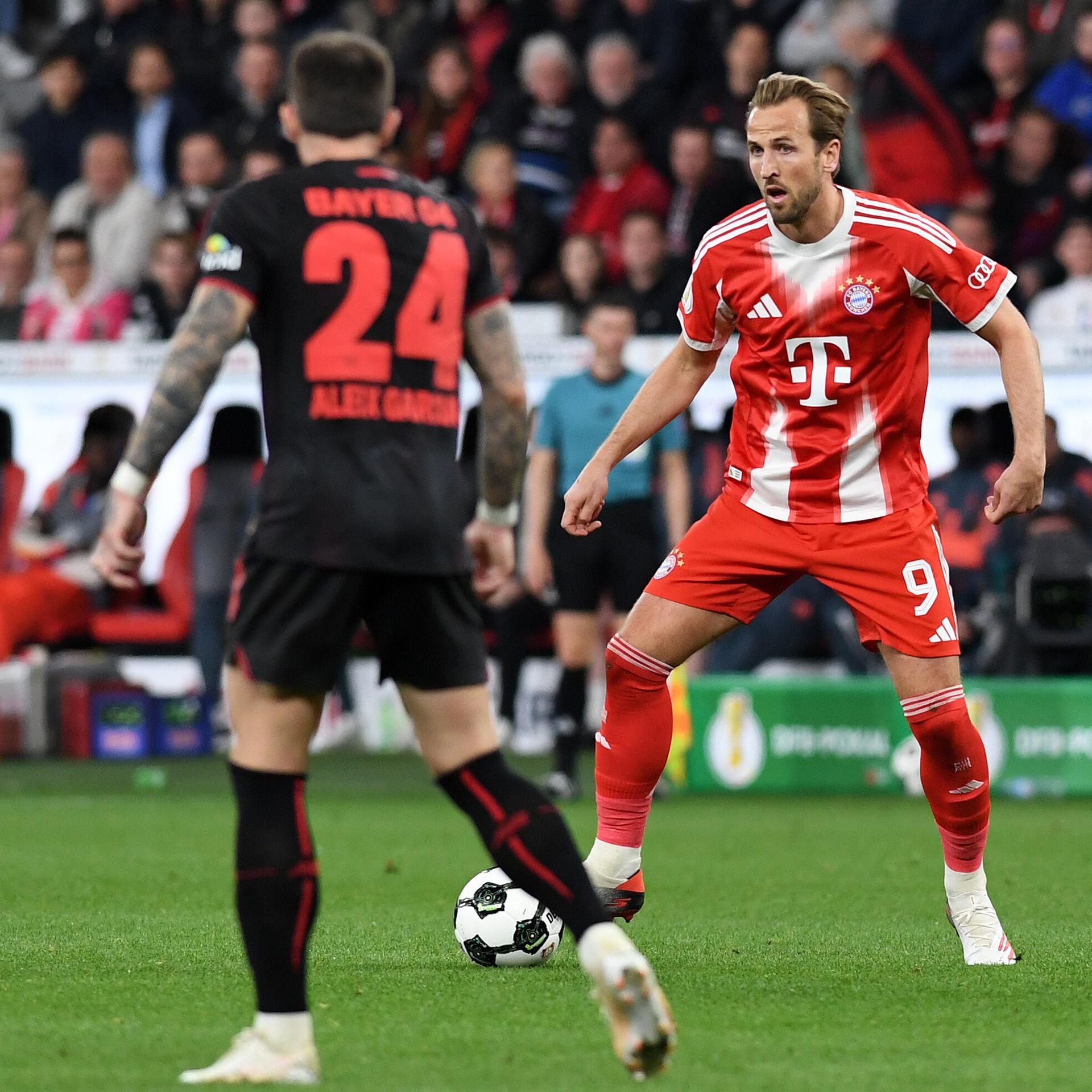 Kane geht mit dem Ball am Fuß auf einen Gegenspieler zu. Kane trägt ein rotes Trikot, der Gegenspieler spielt in schwarz. Im Hintergrund steht Bayern-Trainer Vincent Kompany.
