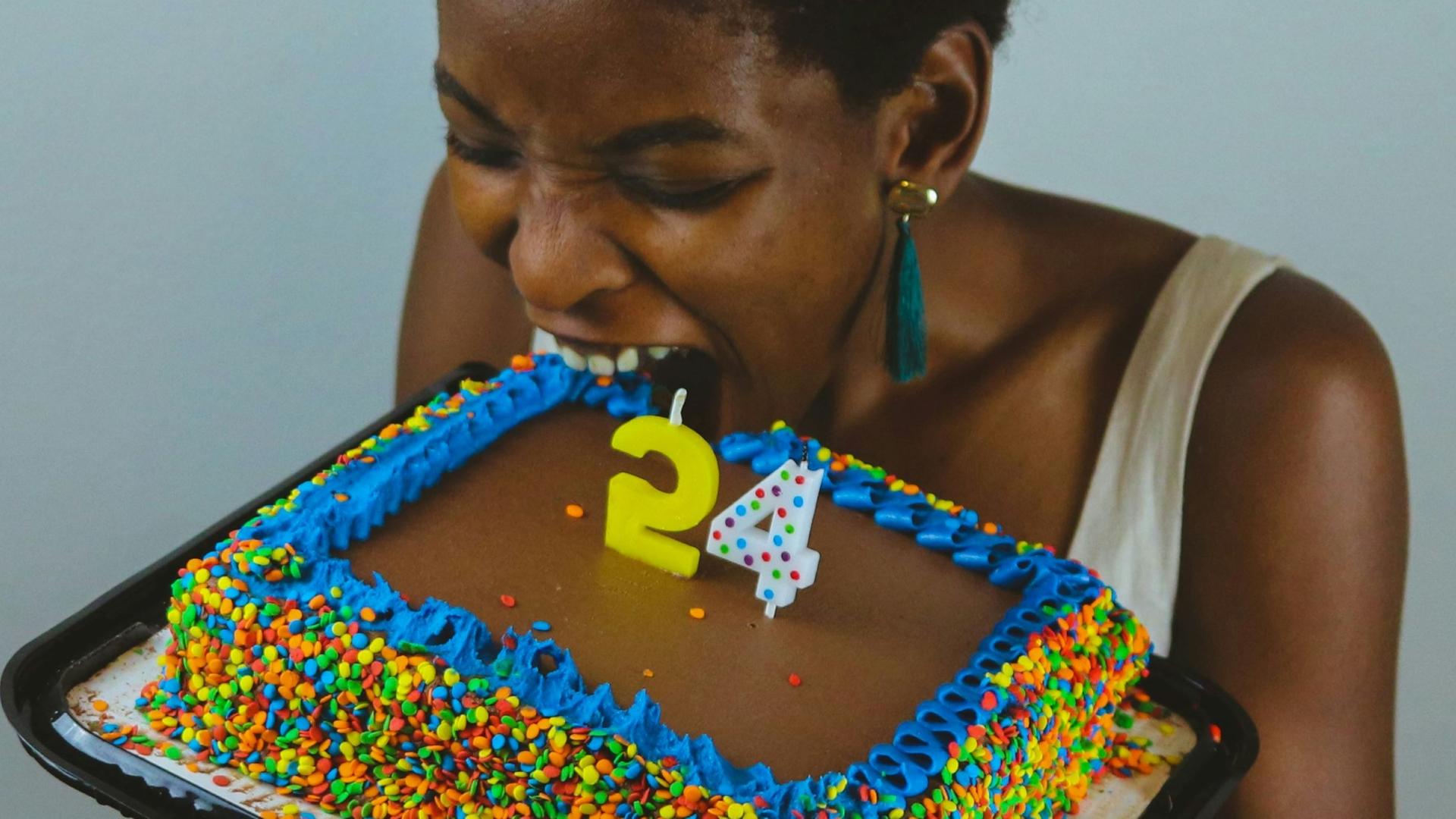 Eine Frau beißt genussvoll in einen mit bunten Zuckerperlen verzierten Geburtstagskuchen, auf dem zwei Kerzen aufgestellt sind, die die Ziffer "24" ergeben.