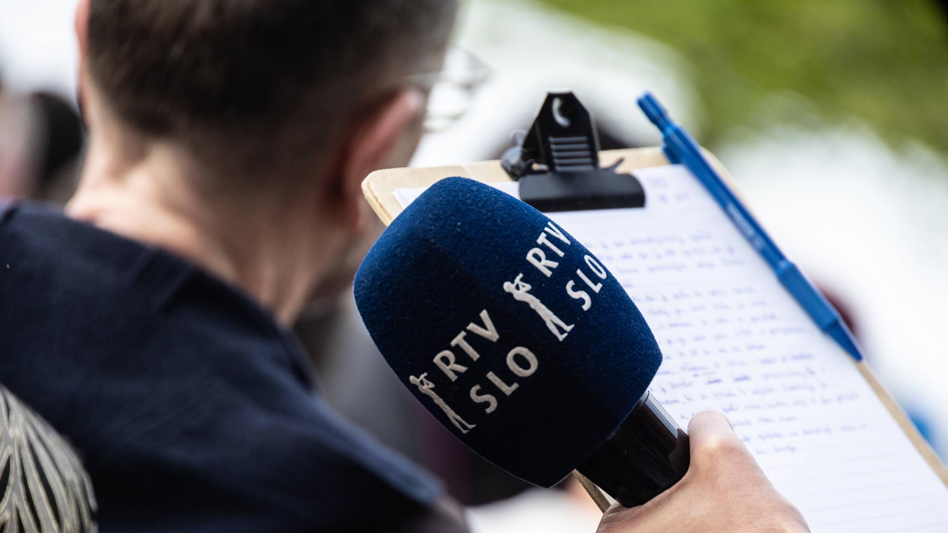 Ein Mikrofon von RTV Slovenija (Radio-Fernsehen Slowenien). Dahinter verschwommen ein männlicher Kopf von hinten und ein Notizblock mit handgeschriebenen Notizen und ein Kugelschreiber