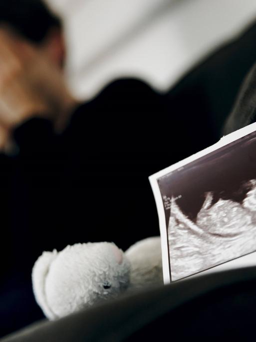 Symbolfoto: Gestellte Aufnahme zum Thema Fehlgeburt. Das Ultraschallbild eines ungeborenen Kindes steht neben einem Kuscheltier waehrend eine Frau im Hintergrund ihr Gesicht in ihren Haenden verbirgt. 