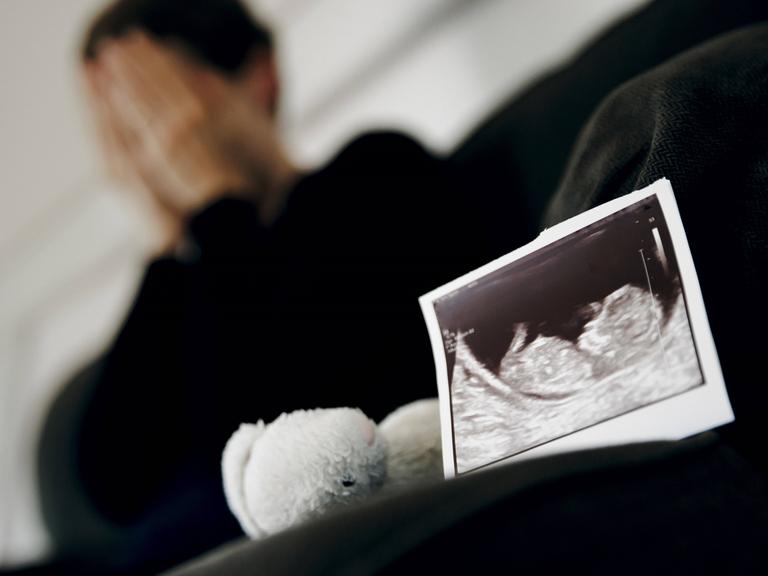 Symbolfoto: Gestellte Aufnahme zum Thema Fehlgeburt. Das Ultraschallbild eines ungeborenen Kindes steht neben einem Kuscheltier waehrend eine Frau im Hintergrund ihr Gesicht in ihren Haenden verbirgt. 