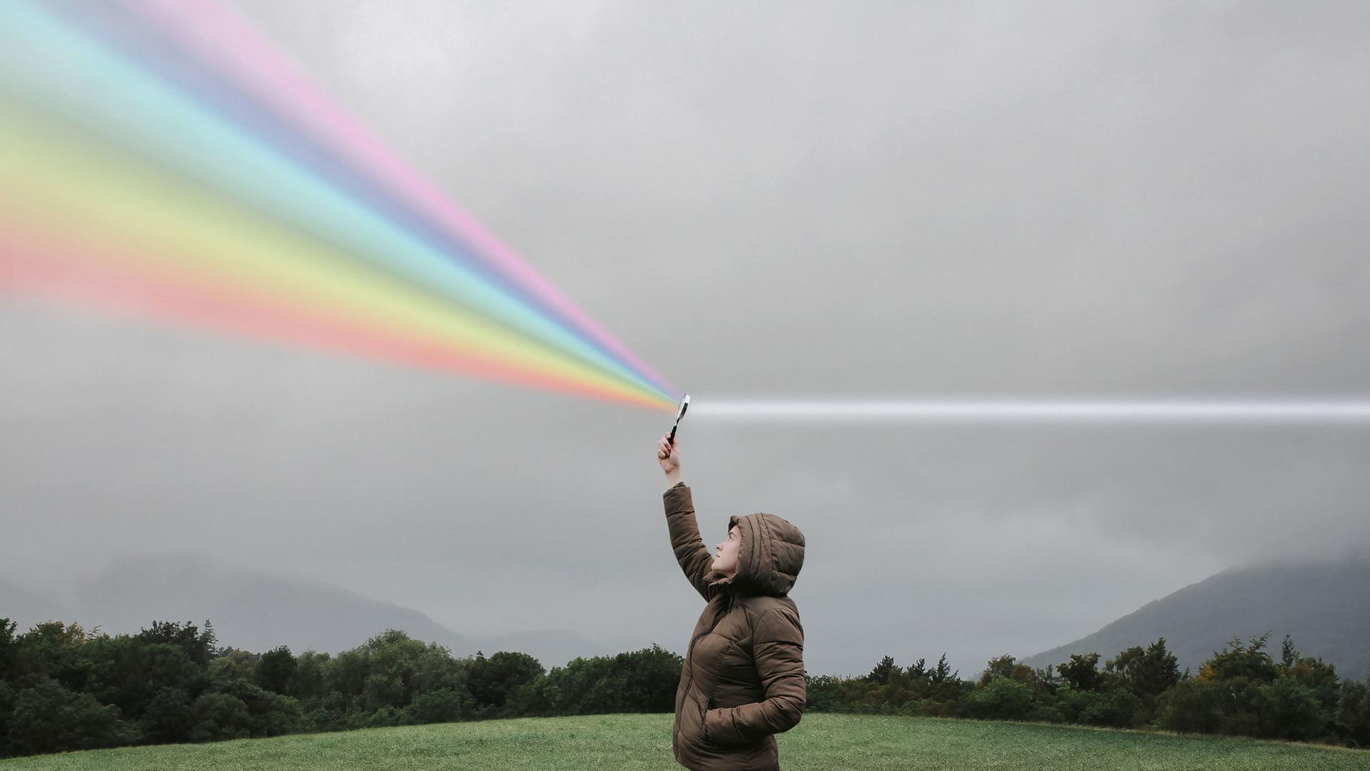 Eine Person steht auf einer Wiese und hält eine Lupe in die Luft in der weißes Licht zum Regenbogenspektrum gebrochen wird.