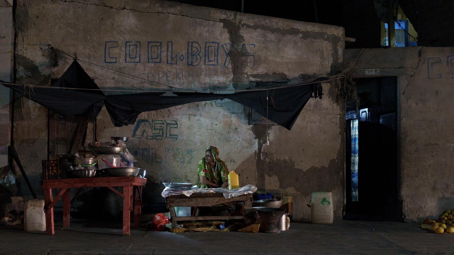 Nachtszene mit einer Straßenverkäuferin, deren Stand von einer kleinen Lampe beleuchtet wird.