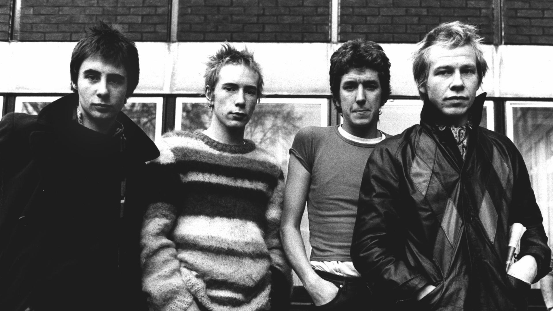 Schwarz-Weiß-Aufnahme der Band Sex Pistols, die nebeneinander vor einem Haus stehen, Hände in den Hosentaschen.