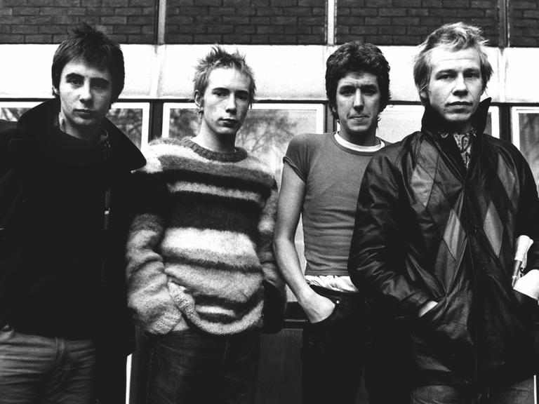 Schwarz-Weiß-Aufnahme der Band Sex Pistols, die nebeneinander vor einem Haus stehen, Hände in den Hosentaschen.