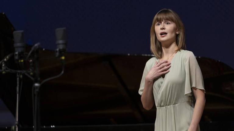 Olga Surikova singt auf einer dunklen Bühne in einem schlichten hellen Kleid. 