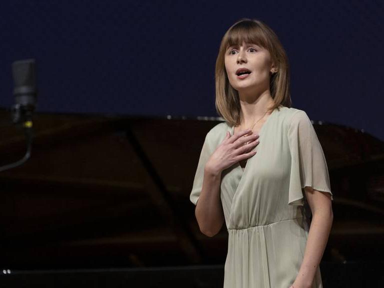 Olga Surikova singt auf einer dunklen Bühne in einem schlichten hellen Kleid. 