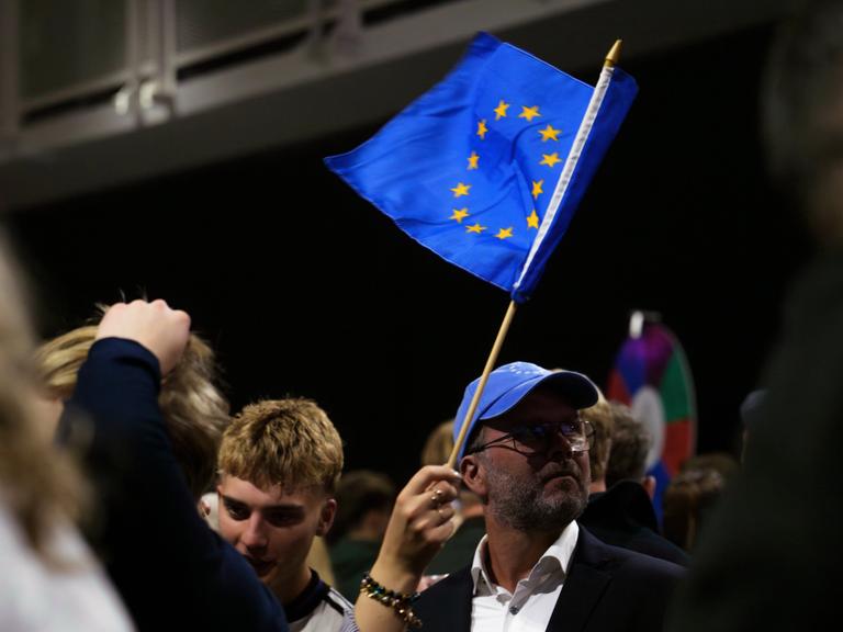 Eine Veranstaltung von Unterstützern für den Beginn von Beitrittsgesprächen mit der EU. Menschen stehen zusammen. Eine Person schwenkt eine blaue EU-Fahne mit den gelben Sternen. Eine Veranstaltung von Unterstützern für den Beginn von Beitrittsgesprächen mit der EU. Menschen stehen zusammen. Eine Person schwenkt eine blaue EU-Fahne mit den gelben Sternen.