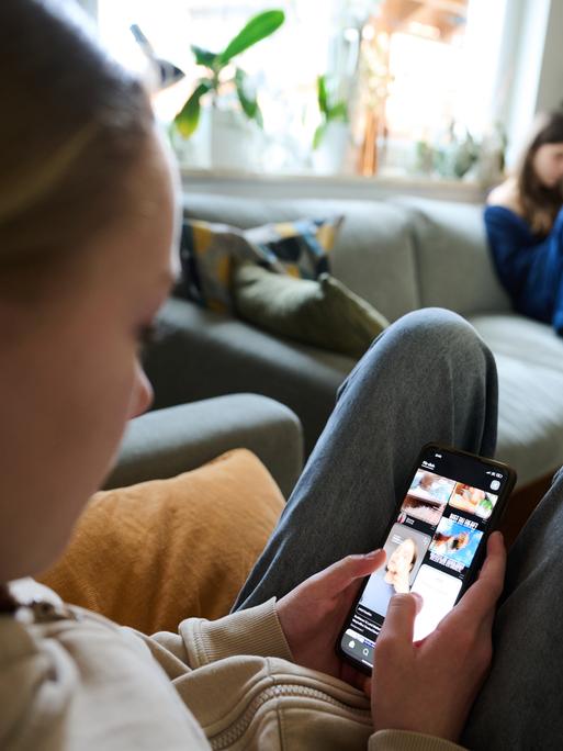 Ein Mädchen nutzt ihr Smartphone, während im Hintergrund ein anderes Mädchen ein Buch liest. (gestellte Szene)