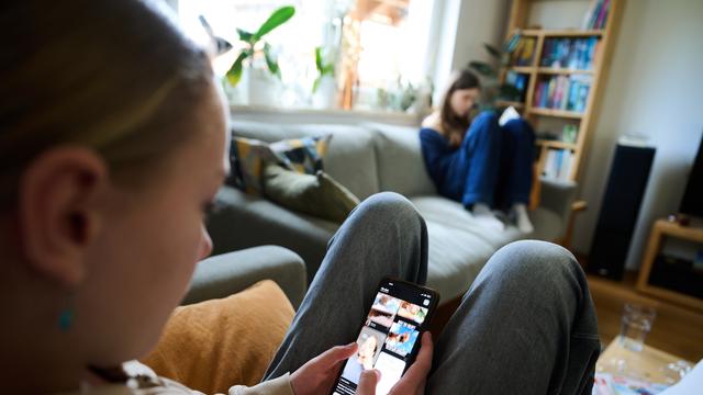 Ein Mädchen nutzt ihr Smartphone, während im Hintergrund ein anderes Mädchen ein Buch liest. (gestellte Szene)
