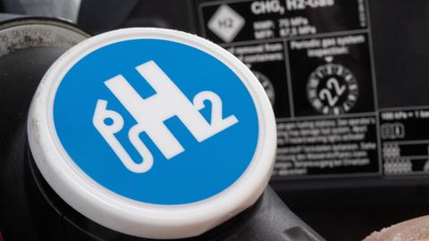 in Mann hält den Zapfhahn einer Wasserstofftankstelle an den Stutzen eines Wasserstoffautos. Auf dem Zahpfhan steht das Symbol "H2".