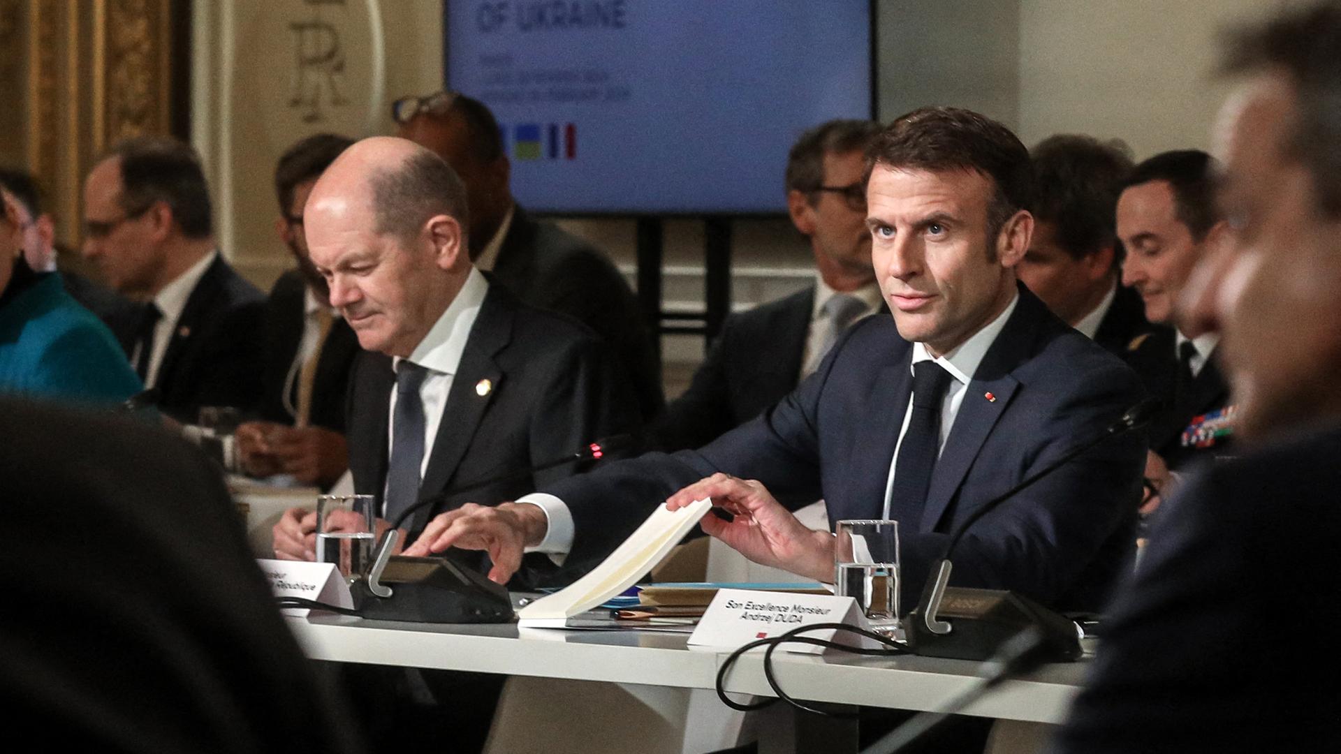 Frankreichs Präsident Emmanuel Macron (r.) und Bundeskanzler Olaf Scholz sitzen nebeneinander an einem Konferenztisch. Daneben und im Hintergrund weitere Personen.