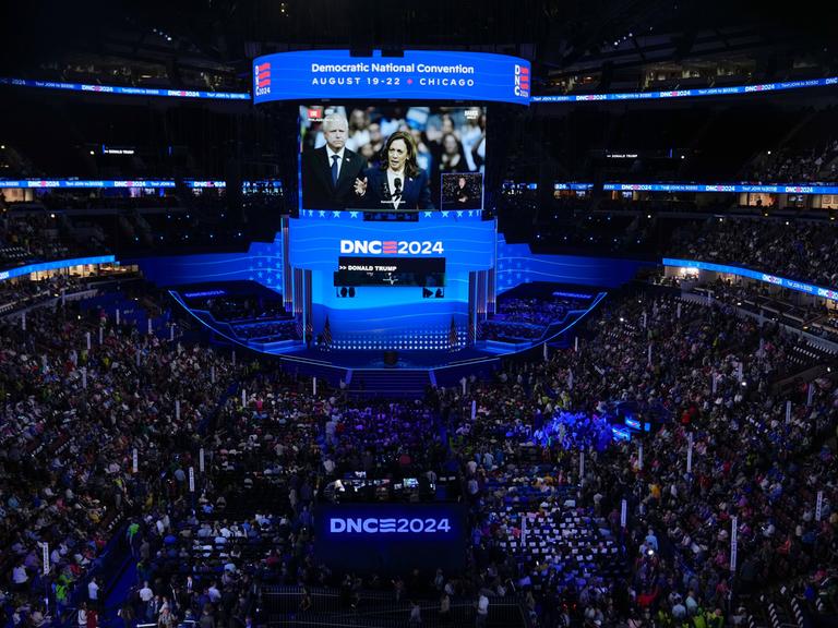 Der volle Saal beim Parteitag der Demokraten eingetaucht in blaues Licht. In der Mitte eine riesige Leinwand, auf der Harris und ihr Mann zu sehen sind. 