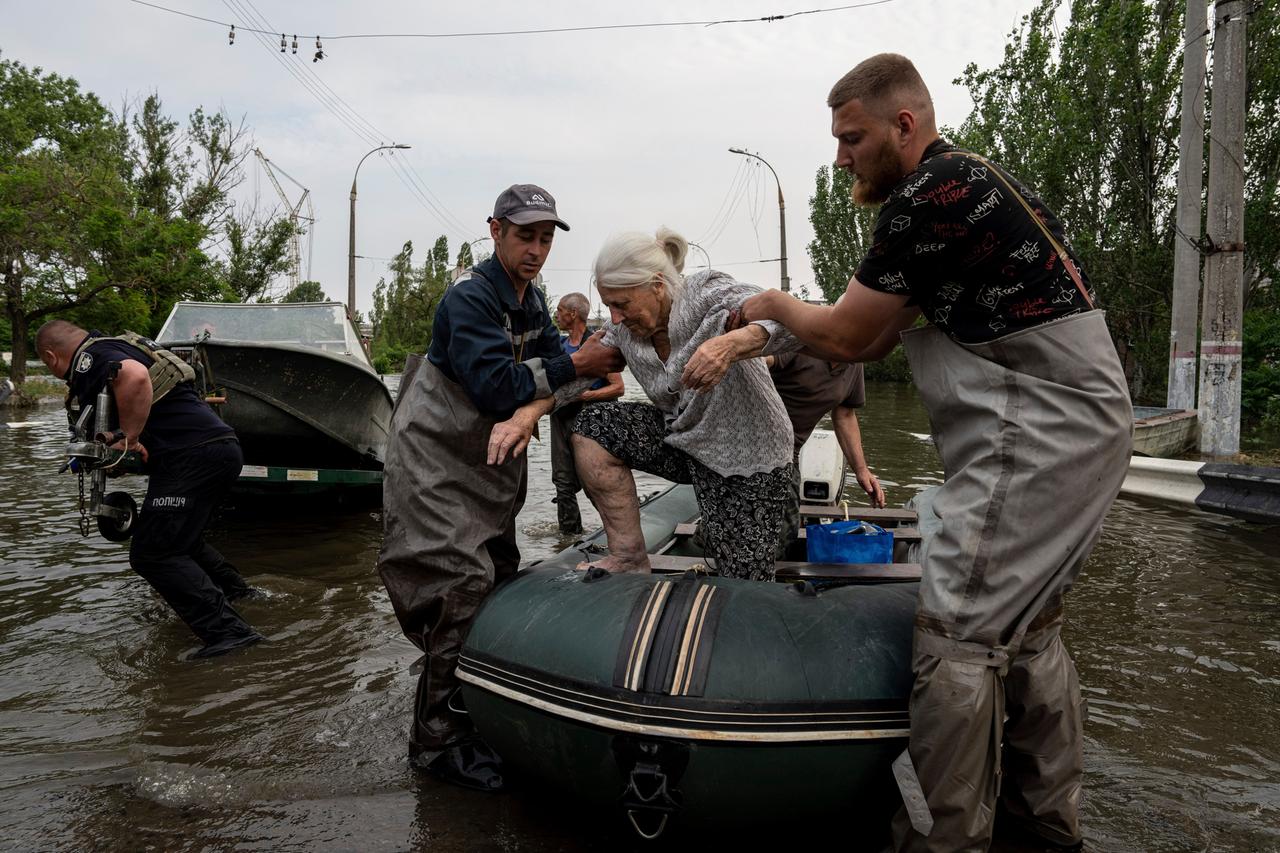 Rettungskräfte evakuieren eine ältere Frau aus einem überfluteten Viertel. Zwei Männer helfen ihr beim Aussteigen aus einem Boot.