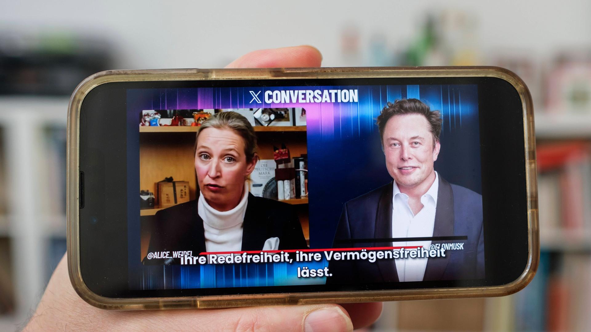 Eine Hand hält ein Mobiltelefon auf dem das Gesprch von Alice Weidel mit Elon Musk auf der Plattform X zu sehen ist. 