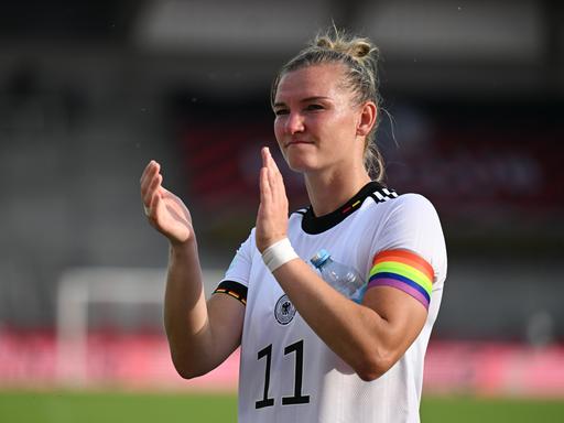 Alexandra Popp steht auf dem Fußballplatz, trägt die Regenbogen-Kapitänsbinde und applaudiert.