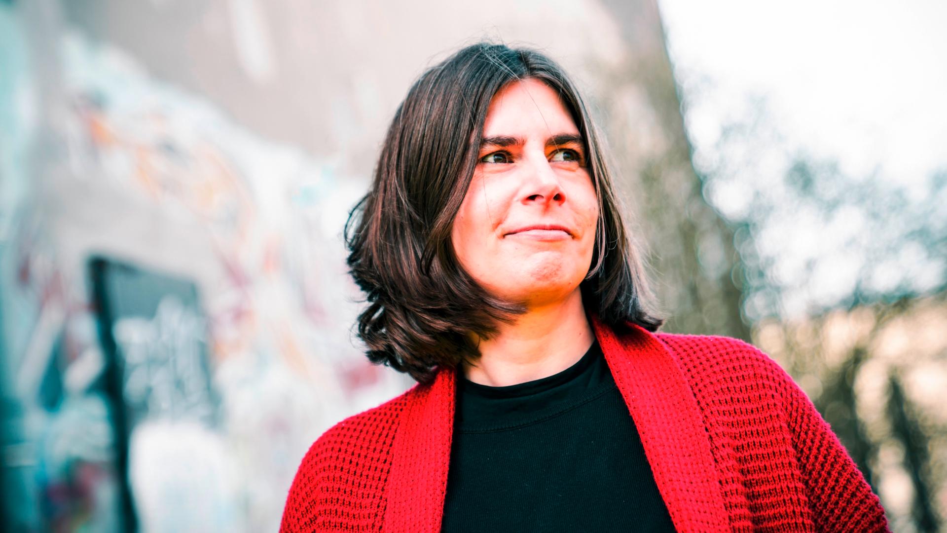 Porträt von Sarah Jäger. Sie hat schulterlanges braunes Haar und trägt eine rote Strickjacke.