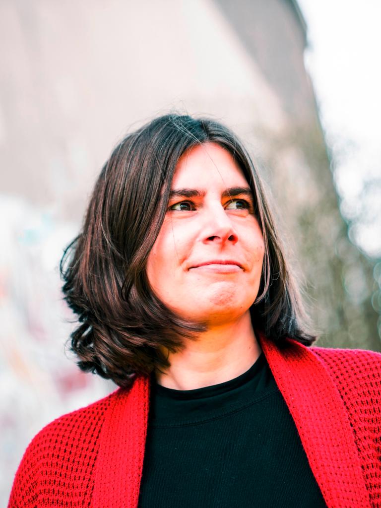 Porträt von Sarah Jäger. Sie hat schulterlanges braunes Haar und trägt eine rote Strickjacke.