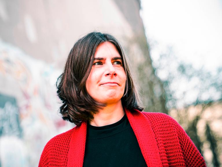 Porträt von Sarah Jäger. Sie hat schulterlanges braunes Haar und trägt eine rote Strickjacke. Porträt von Sarah Jäger. Sie hat schulterlanges braunes Haar und trägt eine rote Strickjacke.