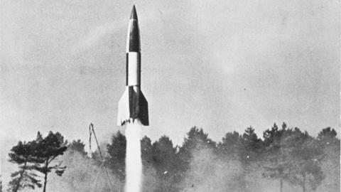 Schwarzweißfoto zeigtt eine Raketvon  deinem bewaldeten Gelände asufsteigen.