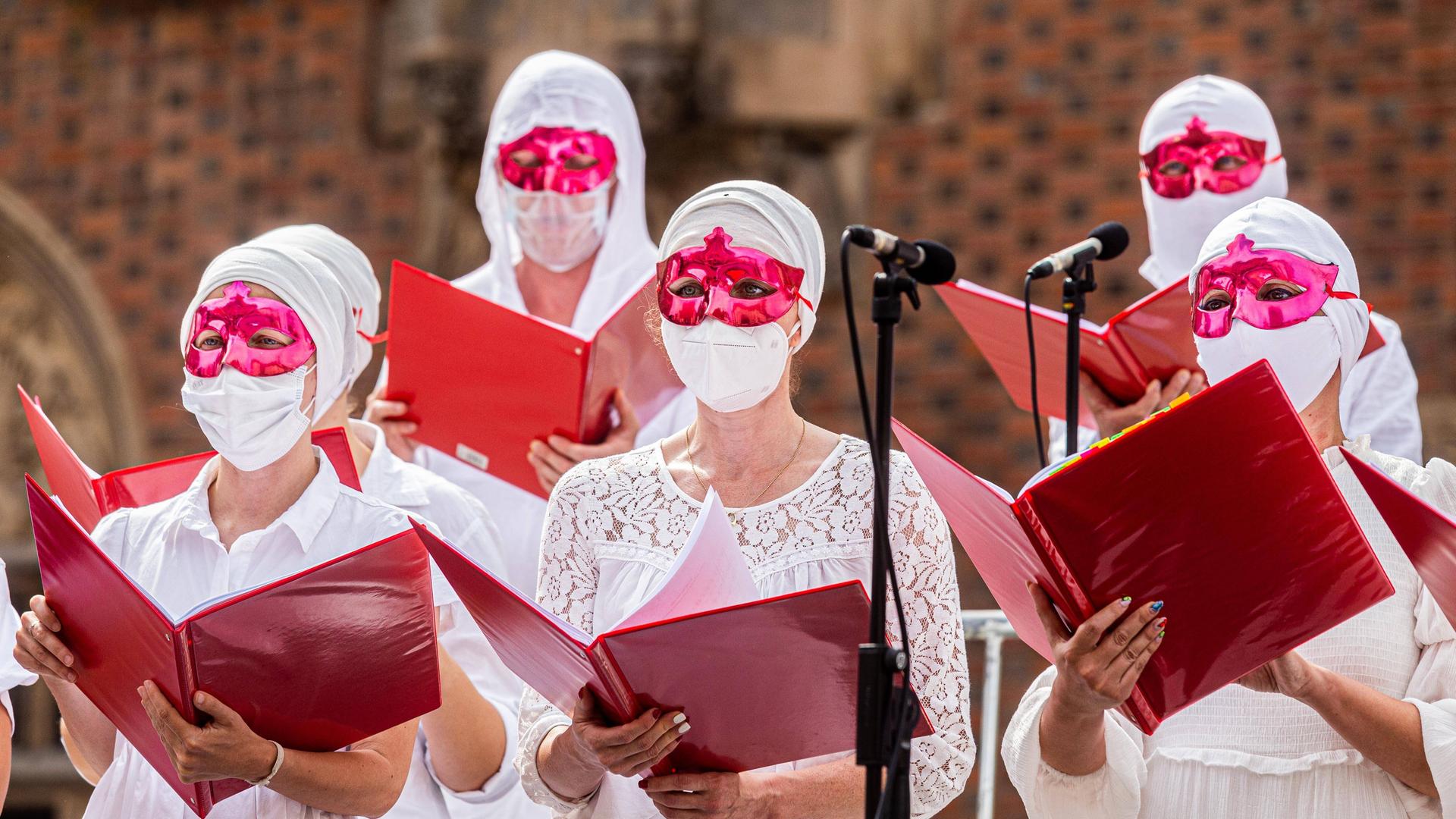 Frauen in weißen Kleidern und weißen, medizinischen Masken und grell roten Augenmasken singen aus großen, roten Mappen.