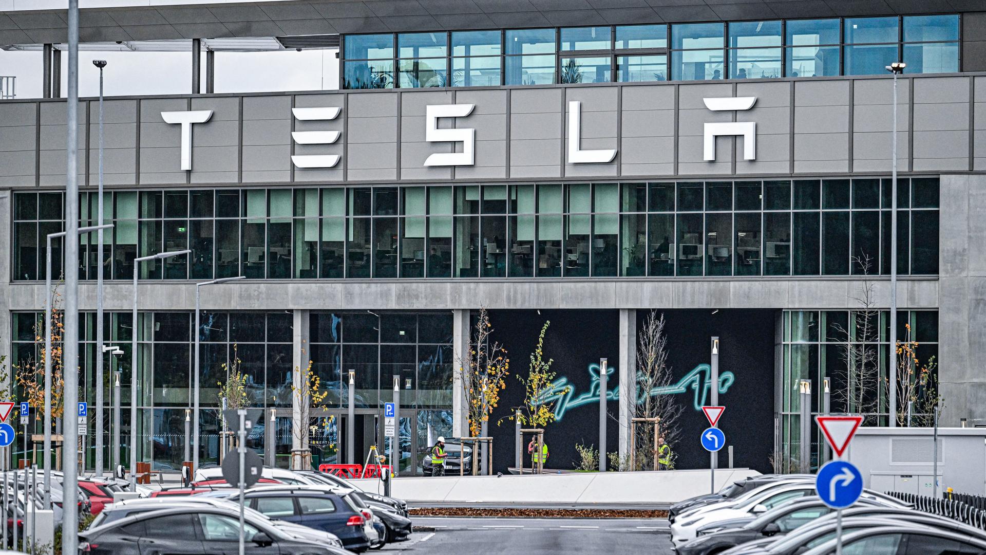 Aufnahme des Hauptgebäudes der Tesla Gigafactory in Grünheide bei Berlin. Im Vordergrund stehen Tesla-Autos auf einem Parkplatz. 