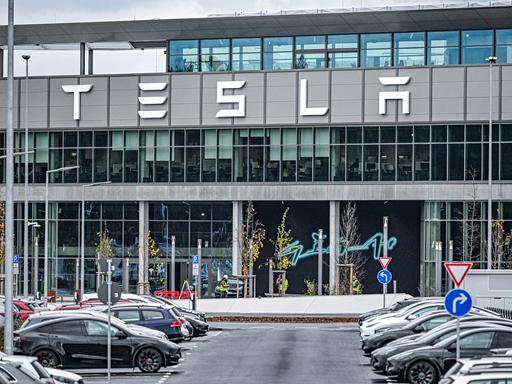Aufnahme des Hauptgebäudes der Tesla Gigafactory in Grünheide bei Berlin. Im Vordergrund stehen Tesla-Autos auf einem Parkplatz. 