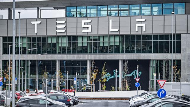 Aufnahme des Hauptgebäudes der Tesla Gigafactory in Grünheide bei Berlin. Im Vordergrund stehen Tesla Autos auf einem Parkplatz.