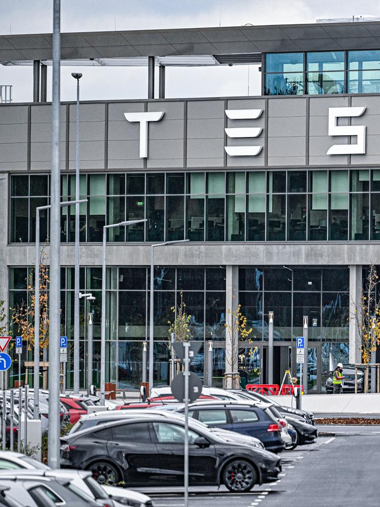 Aufnahme des Hauptgebäudes der Tesla Gigafactory in Grünheide bei Berlin. Im Vordergrund stehen Tesla-Autos auf einem Parkplatz. 