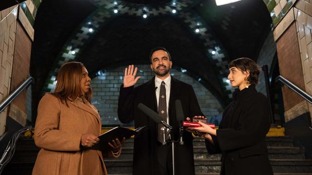 Ein Mann hebt feierlich die rechte Hand, links und rechts von ihm stehen zwei Frauen mit geöffneten Büchern, die sich ihm zuwenden.