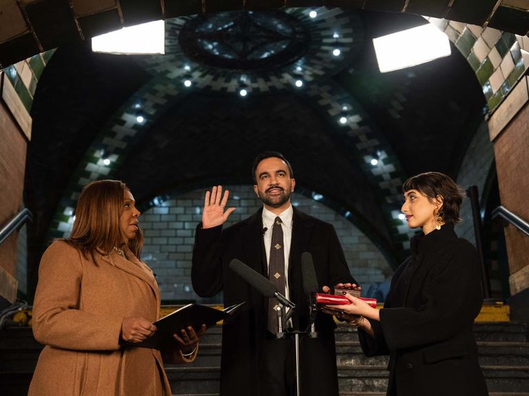 Ein Mann hebt feierlich die rechte Hand, links und rechts von ihm stehen zwei Frauen mit geöffneten Büchern, die sich ihm zuwenden.