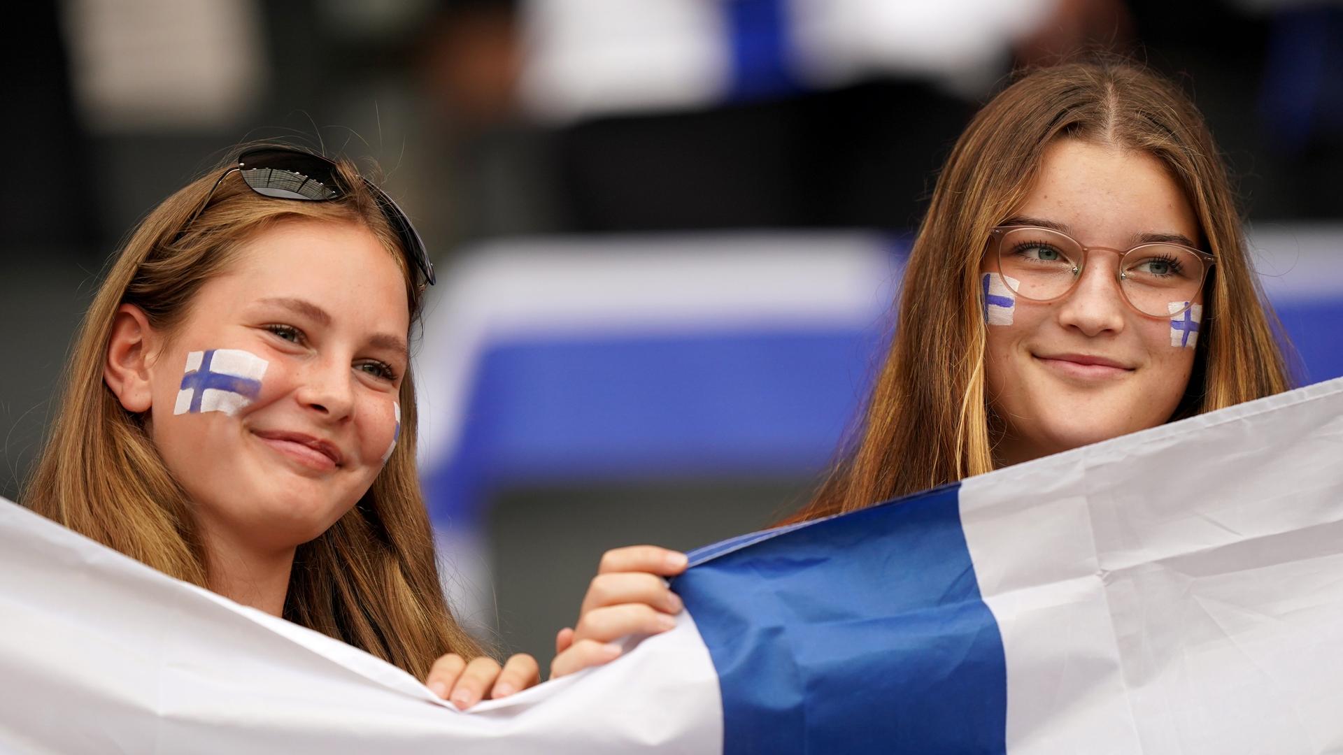 Zwei Finninnen lächeln während eines Fußballspiels. Sie halten eine Finnlad-Flagge und tragen jeweils geschminkte Landesflaggen auf ihren Wangen.