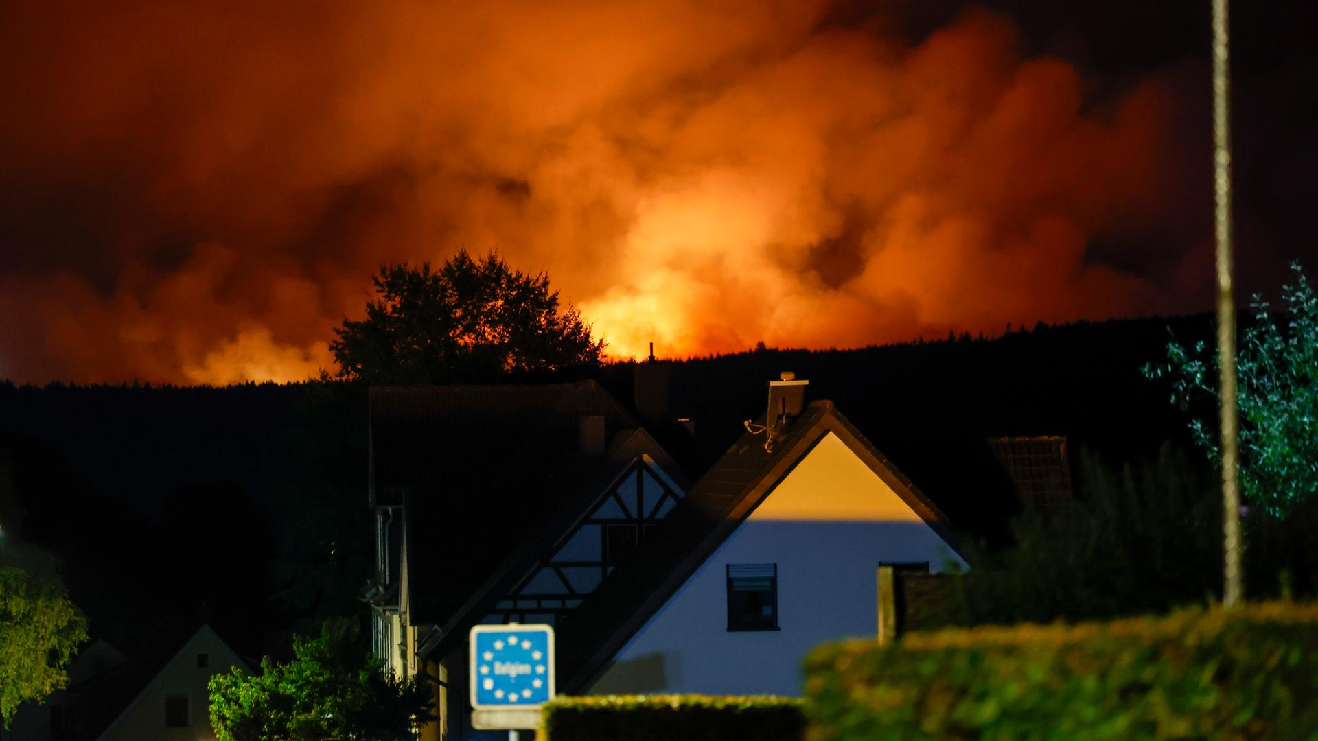 Blick von dem Ortsteil Monschau-Kalterherberg auf die belgische Seite, wo das Hohe Venn in Flammen steht. Der Himmel glüht von den Flammen, im Vordergrund das Schild Belgien und ein Haus. 