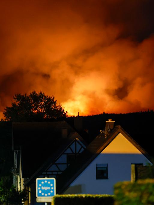 Blick von dem Ortsteil Monschau-Kalterherberg auf die belgische Seite, wo das Hohe Venn in Flammen steht. Der Himmel glüht von den Flammen, im Vordergrund das Schild Belgien und ein Haus. Blick von dem Ortsteil Monschau-Kalterherberg auf die belgische Seite, wo das Hohe Venn in Flammen steht. Der Himmel glüht von den Flammen, im Vordergrund das Schild Belgien und ein Haus.
