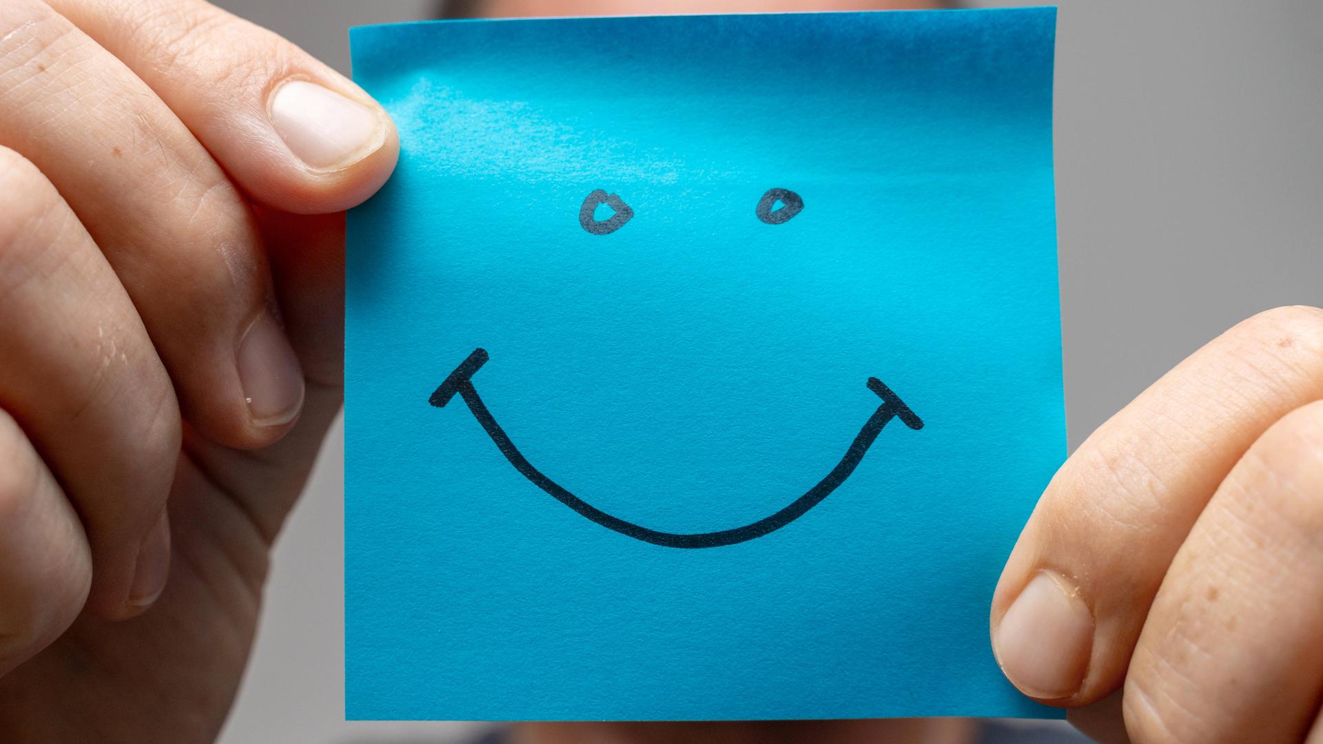 Ein Mann hält mit beiden Händen ein blaues quadratisches Papier auf dem ist ein Smiley gezeichnet