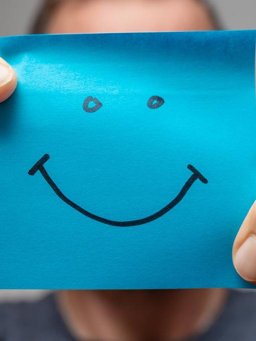 Ein Mann hält mit beiden Händen ein blaues quadratisches Papier auf dem ist ein Smiley gezeichnet