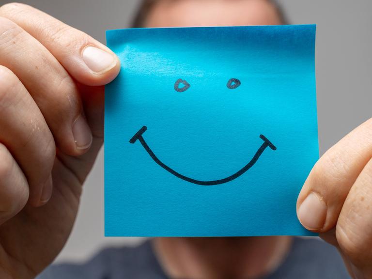 Ein Mann hält mit beiden Händen ein blaues quadratisches Papier auf dem ist ein Smiley gezeichnet